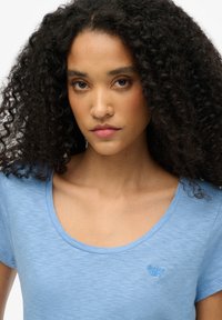 Jasnoniebieska koszulka z krótkim rękawem o okrągłym dekolcie, wykonana z miękkiego materiału, z delikatnie haftowanym logo na piersi.