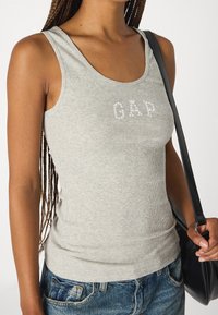 Top de tirantes gris con ribetes que presenta el logo "GAP" en piedras brillantes, combinado con pantalones cortos de mezclilla y un bolso negro de hombro.
