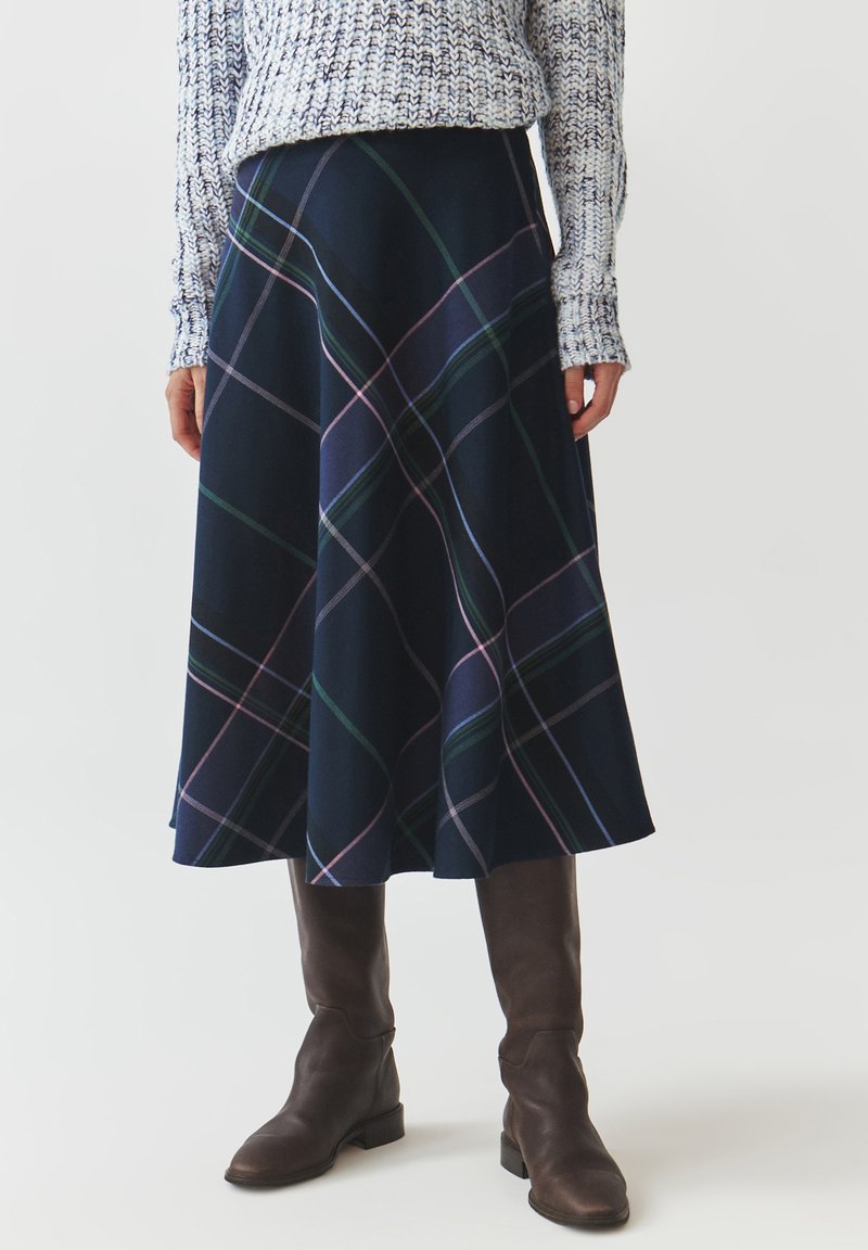 Jupe A-line à carreaux navy avec des lignes vertes et roses, fabriquée en matériau doux. Associée à des bottes marron jusqu'au genou. Pull en tricot texturé au-dessus.