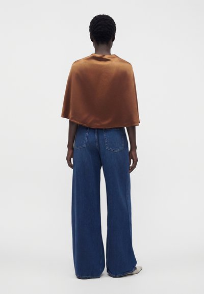 Personne aux cheveux courts et bouclés vue de dos portant un petit capelet en satin marron et un jean bleu à jambes larges avec des chaussures blanches.