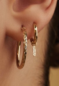 Gold earrings featuring a textured, hammered finish on one hoop and a sleek, polished design on the smaller hoop, with a short pendant accent.