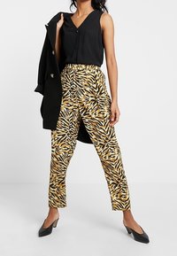 Kalhoty se zvířecím potiskem v žluté a černé barvě, vysoký pas, spárované s černým bezrukávovým topem a kabátem, a černými podpatky.