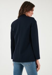 Mornarsko modra krojena blazer jakna z gladkim materialom, zareznimi reverji in prilegajočo silhueto. Ima srednji šiv na hrbtu in dolge rokave.