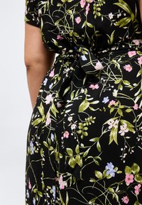 Μαύρο φόρεμα με λουλουδάτο μοτίβο σε ροζ, μπλε και πράσινες βοτανικές αποχρώσεις, με ζώνη δεμένη στη μέση και κοντά μανίκια σε ένα άτομο.