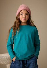 Türkisfarbenes Langarmshirt aus weicher Baumwolle. Kombiniert mit lässiger Jeans trägt das Mädchen eine pinke Strickmütze und zeigt einen entspannten, alltäglichen Stil.