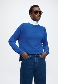 Kék pulóver kötött anyagból, kerek nyakkivágással, raglán ujjakkal és egy kis fekete logóval a mellkasnál, magas derekú farmernadrággal párosítva.