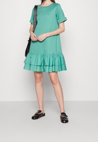 Robe en coton teal avec un col ras du cou, des manches courtes roulées et un ourlet à volants superposés. Portée avec des chaussures noires à enfiler.