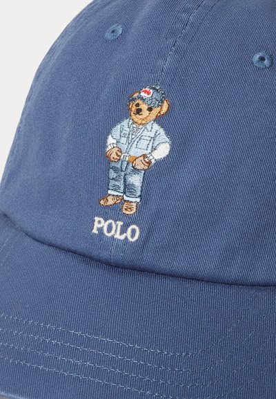 Μπλε μπέιζμπολ καπέλο με κεντημένη αρκούδα που φοράει ντένιμ φόρεμα και καπέλο, στέκεται με τα χέρια στη μέση πάνω από το κείμενο "POLO".