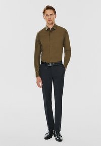 Mężczyzna ubrany w oliwkową koszulę z guzikami, czarny pasek, czarne spodnie eleganckie i czarne buty, stoi z jedną ręką w kieszeni.
