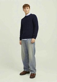 Junger Mann steht da, trägt einen navyblauen Pullover mit Zopfstrickmuster, helle blaue Jeans, braune Schuhe und ein hellblaues Hemd mit Kragen darunter.