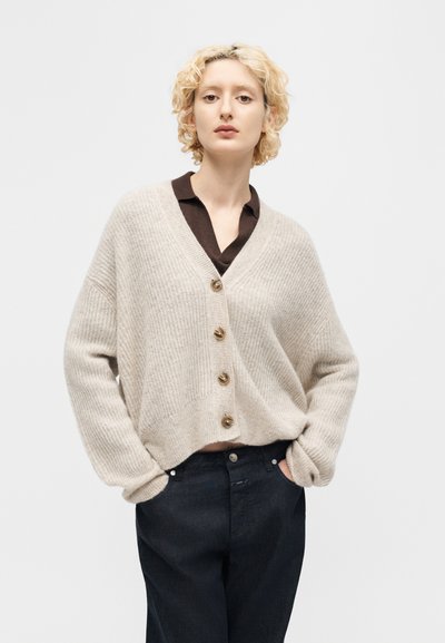 Ξανθό άτομο που φοράει μπεζ ζακέτα με κουμπιά πάνω από σκούρο πουκάμισο με γιακά, με τα χέρια στις τσέπες μαύρου παντελονιού, ουδέτερο φόντο.