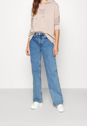 Beige huppari logolla, yhdistettynä vaalean sinisiin leveälahkeisiin farkkuihin. Valkoiset lenkkarit täydentävät asua. Yksinkertainen, rento ilme.