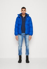 Giacca puffer elettrica di colore blu con design trapuntato, abbinata a una polo navy e jeans azzurri chiari. Stivali neri con lacci.