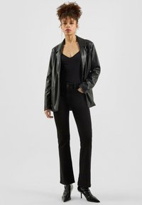 Schwarze Kunstleder-Jacke über einem schwarzen Top, kombiniert mit schwarzen, weit geschnittenen Jeans und spitzen schwarzen Absätzen. Glatte Textur mit minimalen Details.