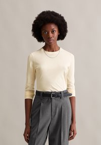 Camisa de manga longa amarelo-claro combinada com calças cinza risca de giz; o conjunto inclui um cinto preto e um colar de corrente prateada.