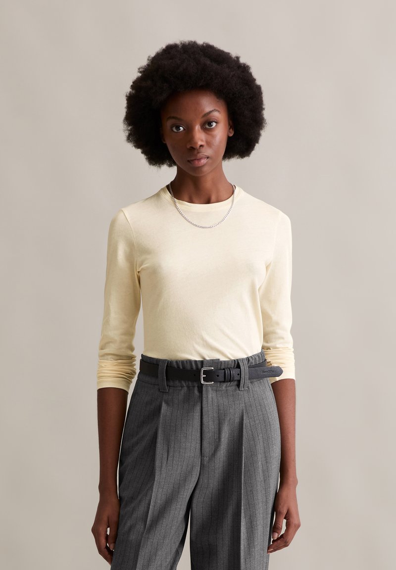 Camisa de manga longa amarelo-claro combinada com calças cinza risca de giz; o conjunto inclui um cinto preto e um colar de corrente prateada.