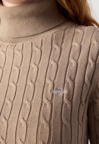 Pull beige à col roulé en maille torsadée avec un petit logo brodé en rouge, blanc et bleu sur la poitrine, porté par une personne aux cheveux longs.