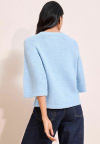 Pulover de un albastru deschis, cu textură moale, cu mâneci trei sferturi și un croi lejer, purtat cu pantaloni din denim închis. Design simplu, aspect general moale.