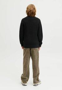 Schwarzer Strickpullover mit strukturierter Oberfläche, kombiniert mit lockeren, hellbraunen Hosen. Robuste Schuhe runden das Outfit ab.