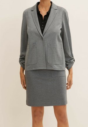 Persoon draagt een grijze blazer met twee voorzakken en een bijpassende grijze rok over een zwart overhemd met knopen, staand tegen een effen achtergrond.