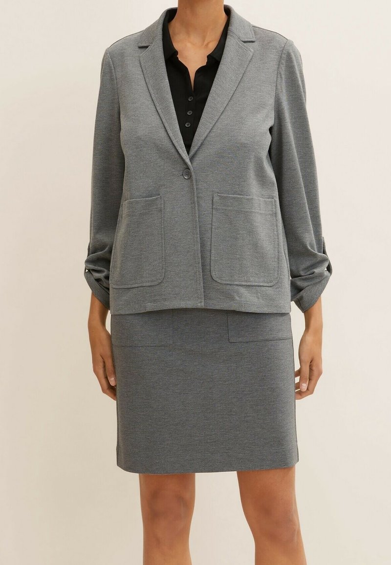 Personne portant un blazer gris avec deux poches avant et une jupe grise assortie sur un haut noir à boutons, debout devant un fond uni.