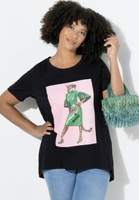 Czarny t-shirt z kolorową grafiką lamparta w okularach przeciwsłonecznych. Modelka łączy go z zieloną torebką z frędzlami i jasnoniebieskimi jeansami.