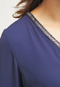 Top blu navy con scollatura a v e decorazioni metalliche lungo il collo, realizzato in un tessuto morbido e leggero.
