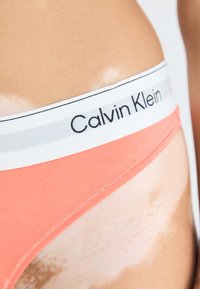 Calvin Klein dames bikini in koraal met een witte tailleband met zwart logo. Gladde stof, aansluitend ontwerp, zichtbare stikdetails.