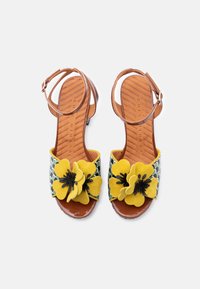 Sandales multicolores avec une bride verte et blanche à motifs ornée de fleurs jaunes, des accents noirs, ainsi qu'une semelle et une bride de cheville marron.