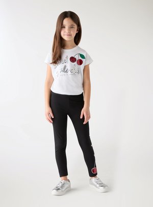 Młoda dziewczyna stoi, ubrana w białą koszulkę z cekinowymi wiśniami oraz czarne legginsy z motywem wiśni przy kostce, srebrne trampki.