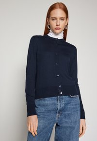 Mornarsko modri pleteni kardigan z okroglim izrezom, zapenjanjem na gumbe in rebrastimi manšetami, oblečen čez belo majico s turtleneck izrezom, v kombinaciji z jeans hlačami.