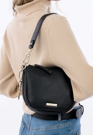 Vrouw draagt een beige geribbelde coltrui en spijkerbroek, en heeft een zwarte leren schoudertas met structuur, gouden ketting en sluitingsdetails.