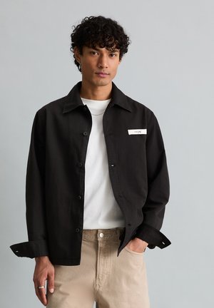 Junger Mann mit lockigem Haar, der eine schwarze, geknöpfte Jacke über einem weißen T-Shirt und beigen Hosen trägt, vor einem schlichten grauen Hintergrund steht.