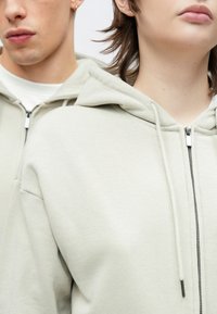 Šviesiai pilkos spalvos džemperiai su kapišonu, pagaminti iš tekstūruotos medžiagos, su priekinėmis užtrauktukais su sidabriniu akcentu ir raišteliais ant kapišono.