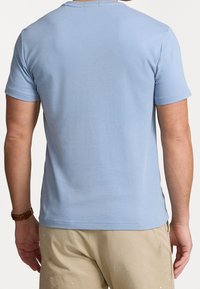 Muž v světle modré krátkosrstvé polokošili a béžových kalhotách, zobrazený zezadu, s hnědými hodinkami na levém zápěstí.