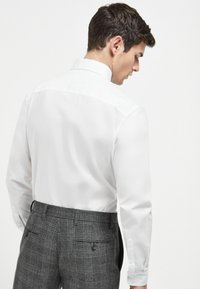 Weißes Langarmhemd mit strukturiertem Kragen, getragen zu grauen Hosen mit Karomuster, die zwei Gesäßtaschen und Knöpfe haben.