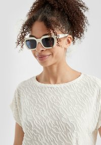 Lunettes de soleil blanches surdimensionnées avec une finition mate, verres teintés foncés, portées avec une chemise crème texturée affichant des motifs ondulés intriqués.