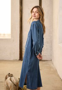 Femme aux longs cheveux blonds portant une robe en denim à manches longues, debout à l'intérieur, regardant par-dessus son épaule avec les mains jointes derrière le dos.