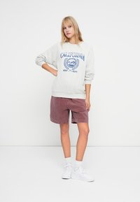 Sweatshirt gris avec un imprimé bleu "California Surf Dept.". Shorts en velours côtelé bordeaux. Des baskets et des chaussettes blanches ajoutent du contraste. Design simpliste.