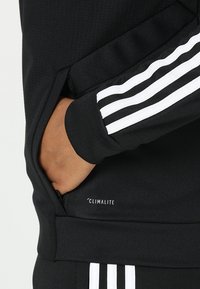 Schwarzer Sportpullover mit weißen Streifen an den Ärmel. Verfügt über eine mit Reißverschluss versehen Tasche und "CLIMATILITE"-Branding. Strukturierter Stoff, lässige Passform.