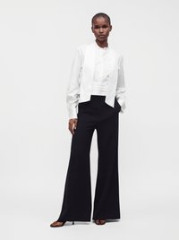 Femme portant une chemise blanche à manches longues et une écharpe, avec un pantalon noir à jambes larges et des sandales noires, debout devant un fond uni.