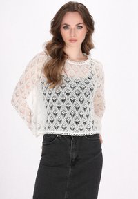 Top bianco in pizzo con design in maglia aperta a motivi, maniche lunghe e orlo a scallop. Indossato sopra una gonna nera, caratterizzata da una silhouette semplice.