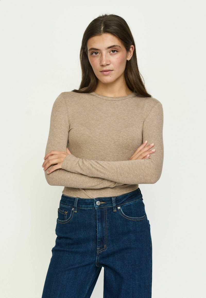 Jeune femme aux longs cheveux bruns portant un haut beige à manches longues et un jean bleu, debout les bras croisés devant un fond uni.