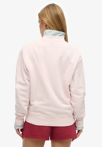 Lichtroze sweatshirt met een hoge kraag, voorzien van een gestreepte blauwe en witte binnenkraag. Draagt losjes met rode shorts voor een ontspannen uitstraling.