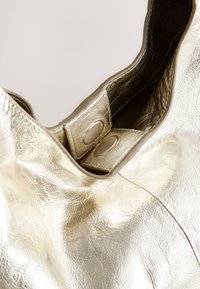 Metalna zlatna torba z teksturirano površino, ki ima mehko obliko in dva gumbna zapiranja na vrhu. Notranja obloga je vidna.