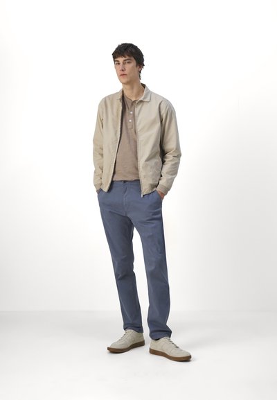 Beige bomberjakke, lys brun polo t-shirt, blå chinos og hvide sneakers. Glat stof, afslappet pasform og diskrete detaljer på jakken.