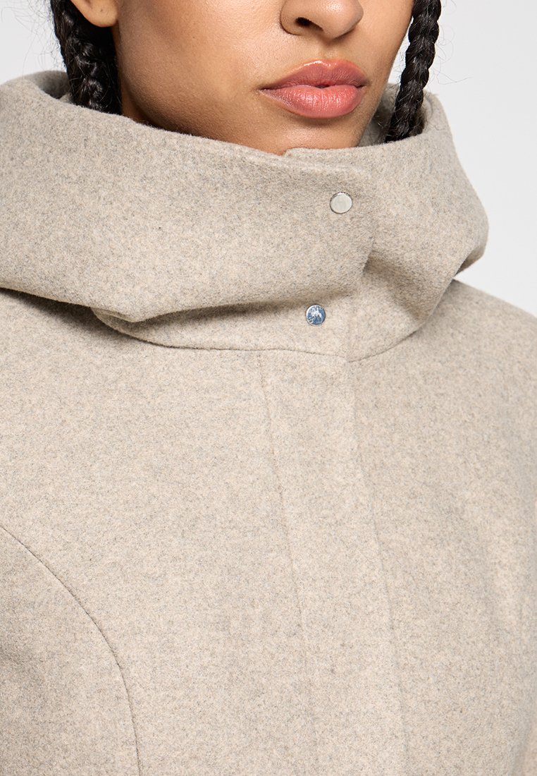 Lichtbeige wollen jas met een hoge kraag, voorzien van drukknoopsluitingen en een textuurafwerking. De stof lijkt zacht en warm te zijn, gemaakt voor comfort.