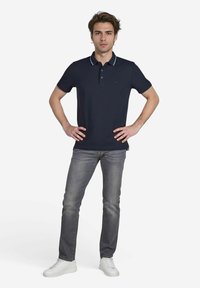 Marineblauw poloshirt met een kraag, witte accenten, korte mouwen, gecombineerd met grijze jeans en witte sneakers, tentoongesteld op een mannelijke model.