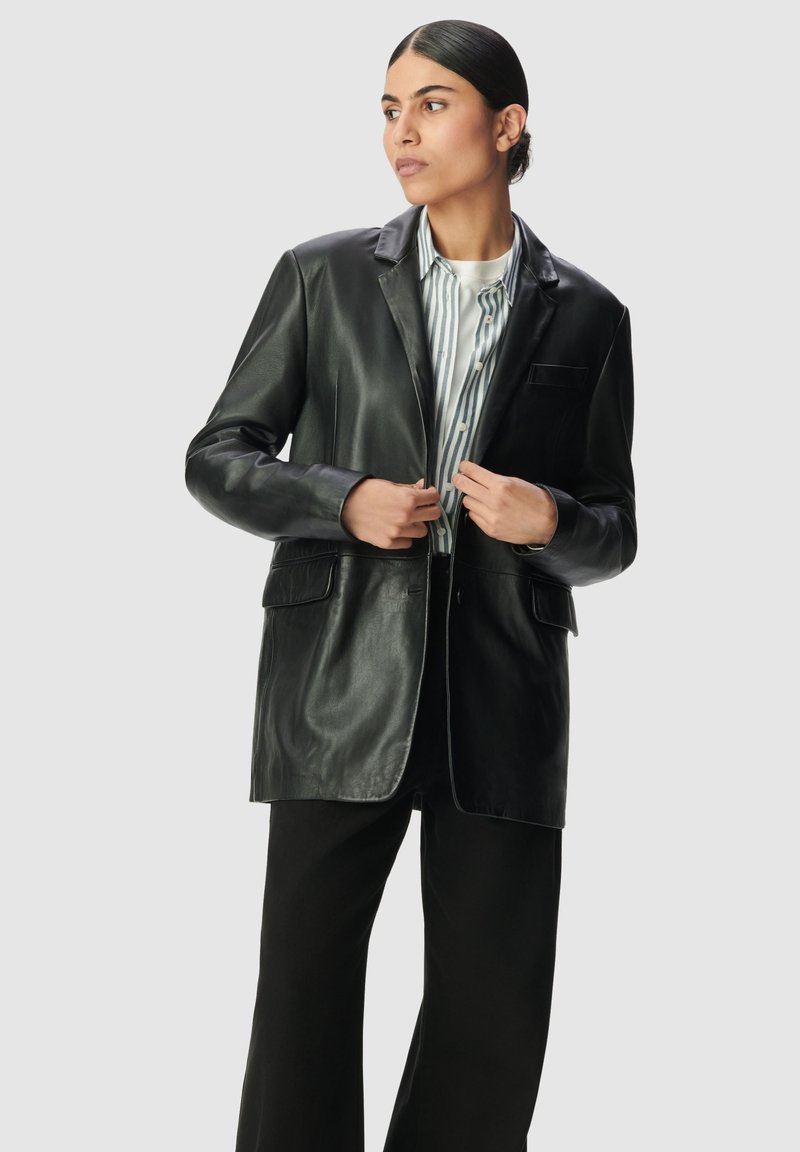 Blazer de cuero negro con solapa muesca, un bolsillo, dos bolsillos frontales y acabado satinado, usado sobre una camisa a rayas y pantalones negros.