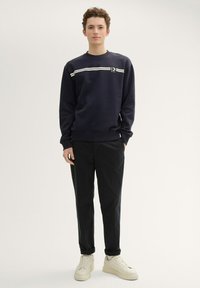 Felpa navy con texture a coste, con una striscia orizzontale bianca e un logo. Abbinata a pantaloni neri e scarpe da ginnastica bianche.